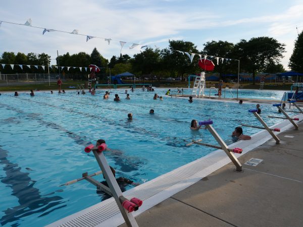 People swim and relax in Middleton’s outdoor public pool while a lifeguard watches. Pool lanes, deck chairs, and a nearby playground are visible, all surrounded by trees under a partly cloudy sky.