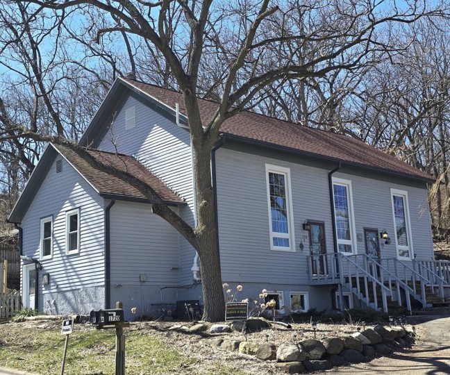 A light blue, single-story house with a brown roof, large windows, a front porch with steps, and leafless trees in the background on a clear day.