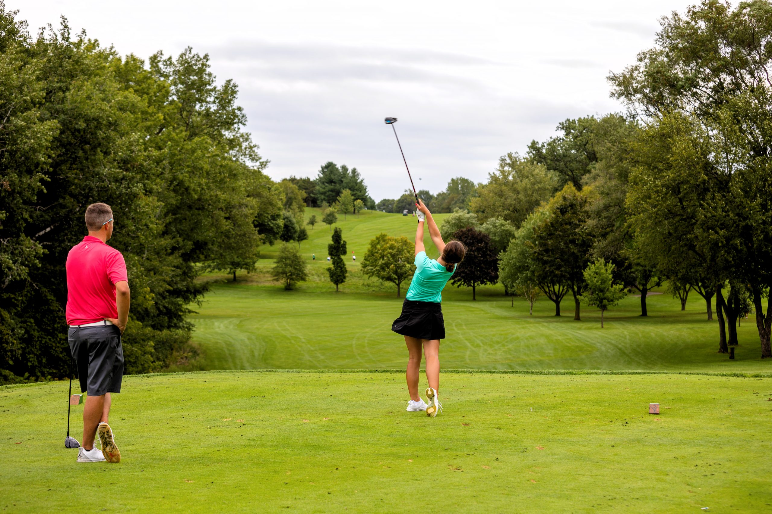 Two people on Pleasant View Golf Course. One, in a teal shirt, swings a golf club while the other, in a red shirt, observes. The course is lush with trees and grass.