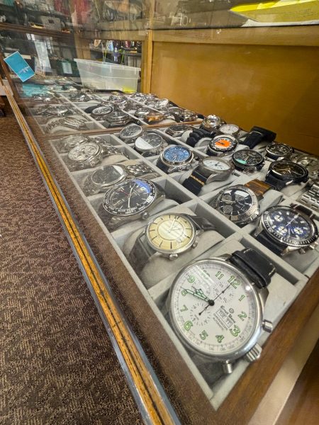 A glass display case containing multiple rows of assorted wristwatches with various styles and bands, set on a beige fabric insert, is showcased in the inviting store environment of University Coin & Jewelry.