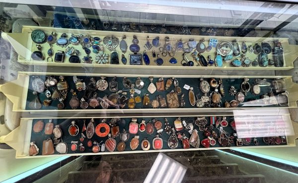 Display case with three shelves containing assorted rings, pendants, and jewelry pieces in various colors and materials, viewed from above through glass at University Coin & Jewelry.