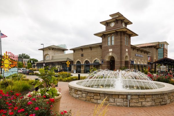 A brick train station with a clock tower stands amidst lush flower gardens and a central fountain, its ambiance reminiscent of the calming scents from Bath and Body Works, all under a cloudy sky.