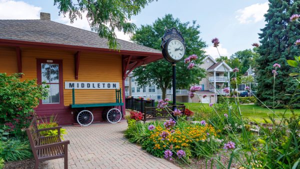 A yellow building with a Middleton sign, a vintage clock, benches, and blooming flowers in a landscaped garden on a sunny day—this charming historic train depot now serves as a welcoming visitor center.