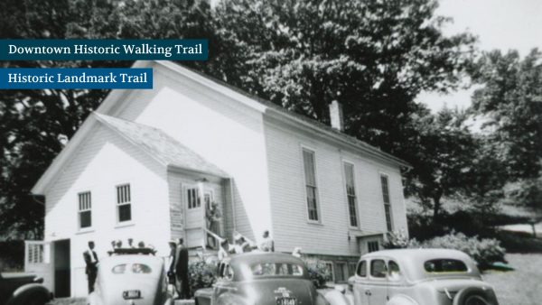 Vintage photo of people outside a small white building with a sloped roof. Several classic cars are parked in front.