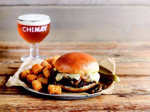 A burger with tater tots and a beer on a plate.