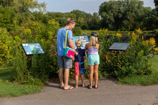 Three children stand on a paved path reading informational signs, surrounded by greenery in Lakeview Park.