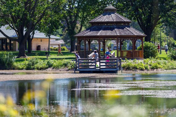 People relax and enjoy the outdoors at a gazebo by a pond in a park.