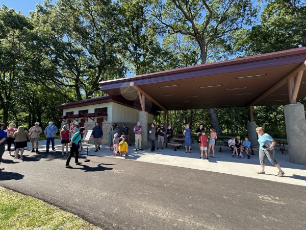A group of people stands and sits under and around a modern pavilion in Middleton Park on a sunny day, with trees and a paved path visible.