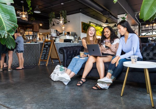 Three women sitting on a couch with laptops in a coffee shop.