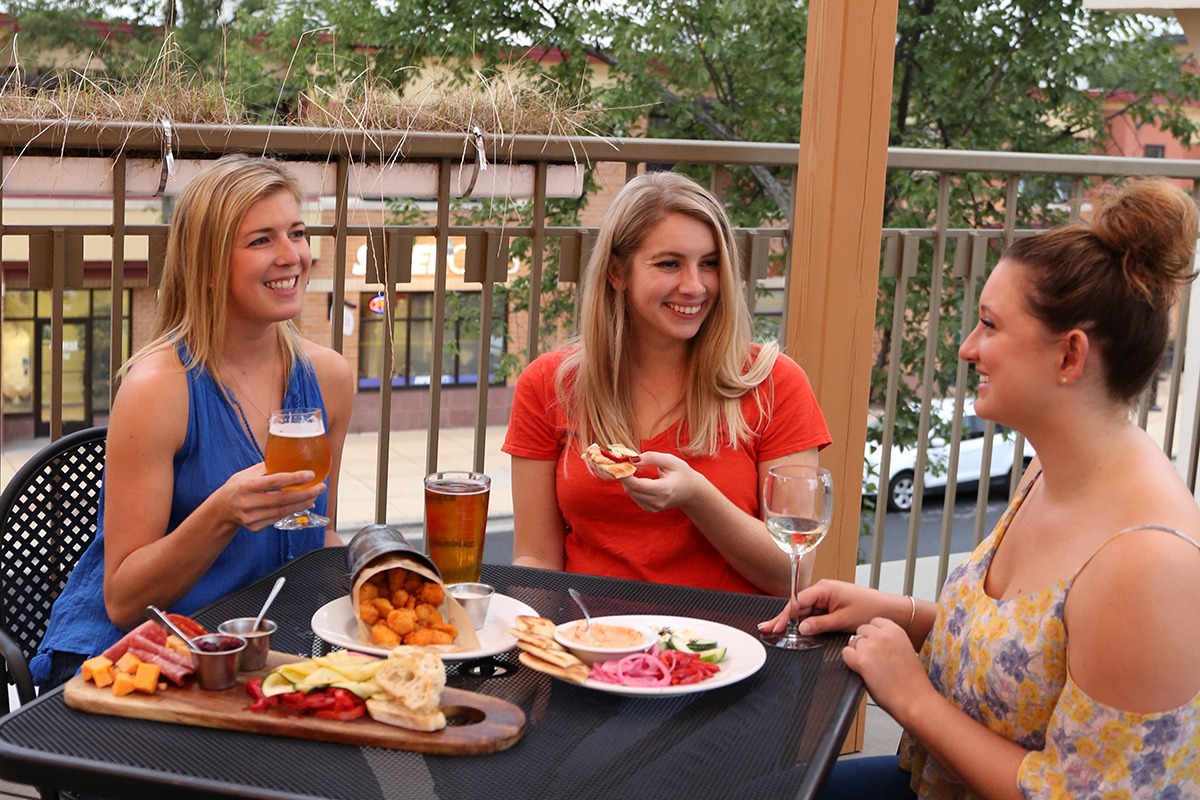 Three women sit at an outdoor dining table enjoying appetizers, beer, and wine, smiling and chatting together on a sunny patio.