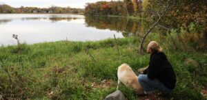 a woman kneeling down next to a dog near a lake