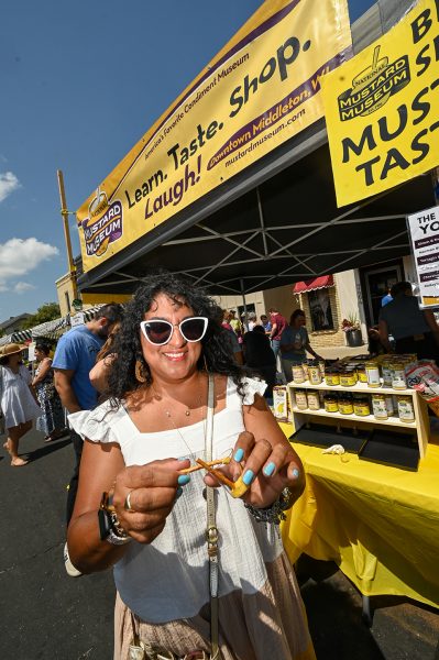 A woman tasting mustards with pretzels beside an outdoor vendor stall during the Mustard Day celebration 2023 in Middleton.
