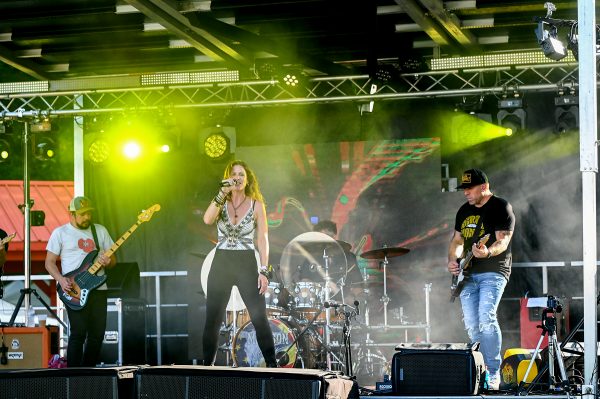 A band performs on an outdoor stage with a female lead singer, a drummer, and two guitarists. Yellow stage lights and smoke effects enhance the scene at the Good Neighbor Festival, turning the live music into a mesmerizing experience.