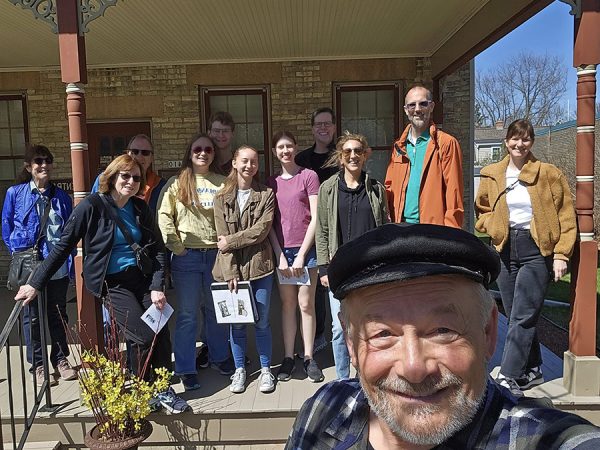 Group of people o the Stroll N Tell, smiling on a sunny day at a historic house entrance, with an older man in a cap, the guide, taking the selfie in the foreground.
