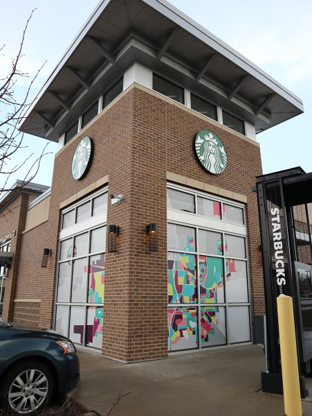 A Starbucks store with a brick exterior, colorful window art, and drive-thru sign visible on a cloudy day.