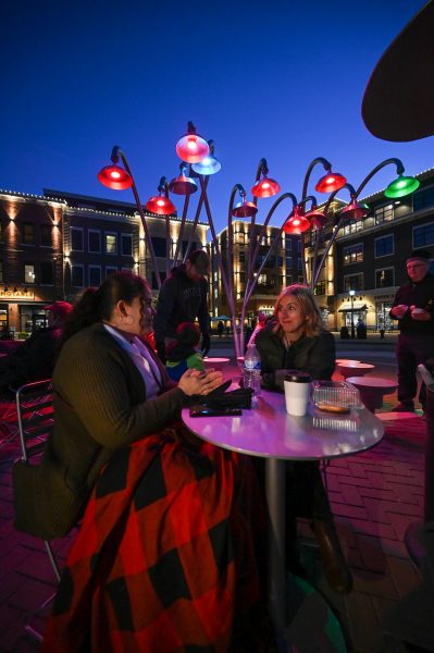Two women sit and talk at an outdoor table in a city plaza at dusk, with colorful overhead lights and buildings illuminated in the background.