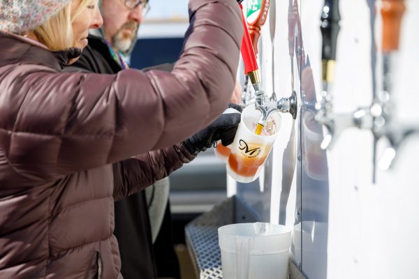 At the bustling winter festival in Middleton, Wisconsin, a person in a brown jacket pours beer from a tap into a clear pitcher at the lively beverage station.