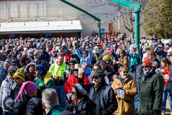 A large crowd of people in winter clothing gathers outdoors at the Bockfest, some holding cups. Strings of lights are hung above, casting a warm glow on the scene, with the Capital Brewery & Bier Garten visible in the background.