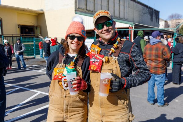 At the Capital Brewery & Bier Garten's winter festival, two people in overalls, adorned with necklaces of snack bags, hold beer mugs at the lively outdoor event. Dressed warmly with hats and sunglasses, they enjoy the festive atmosphere surrounded by fellow attendees.