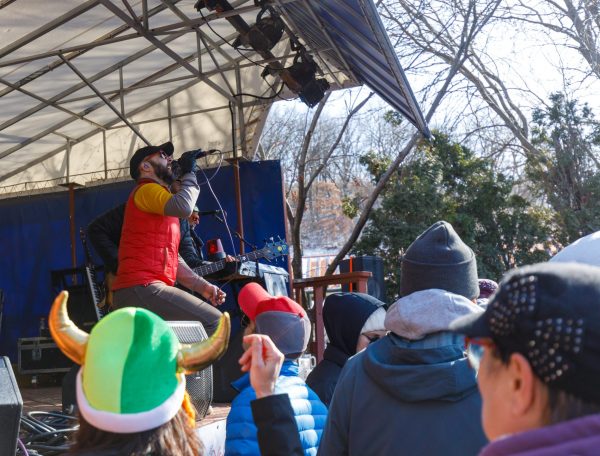 A musician performs on an outdoor stage at Capital Brewery & Bier Garten in Middleton, Wisconsin, holding a microphone. The audience is gathered in front, some wearing hats. Trees and the blue sky of Bockfest are in the background.