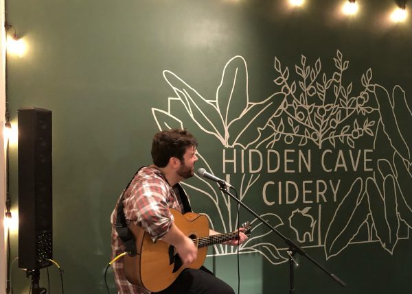 A man playing an acoustic guitar in front of a sign that says hidden cave cidery.