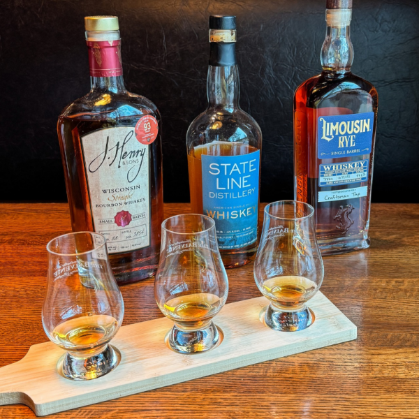 Three bottles of whiskey—J. Henry & Sons, State Line Distillery, and Limousin Rye—arranged behind a wooden tasting flight containing three glasses of amber-colored whiskey.