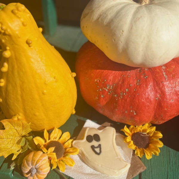A yellow gourd, a stack of white and orange pumpkins, and a ghost-shaped cookie arranged with artificial sunflowers and leaves.