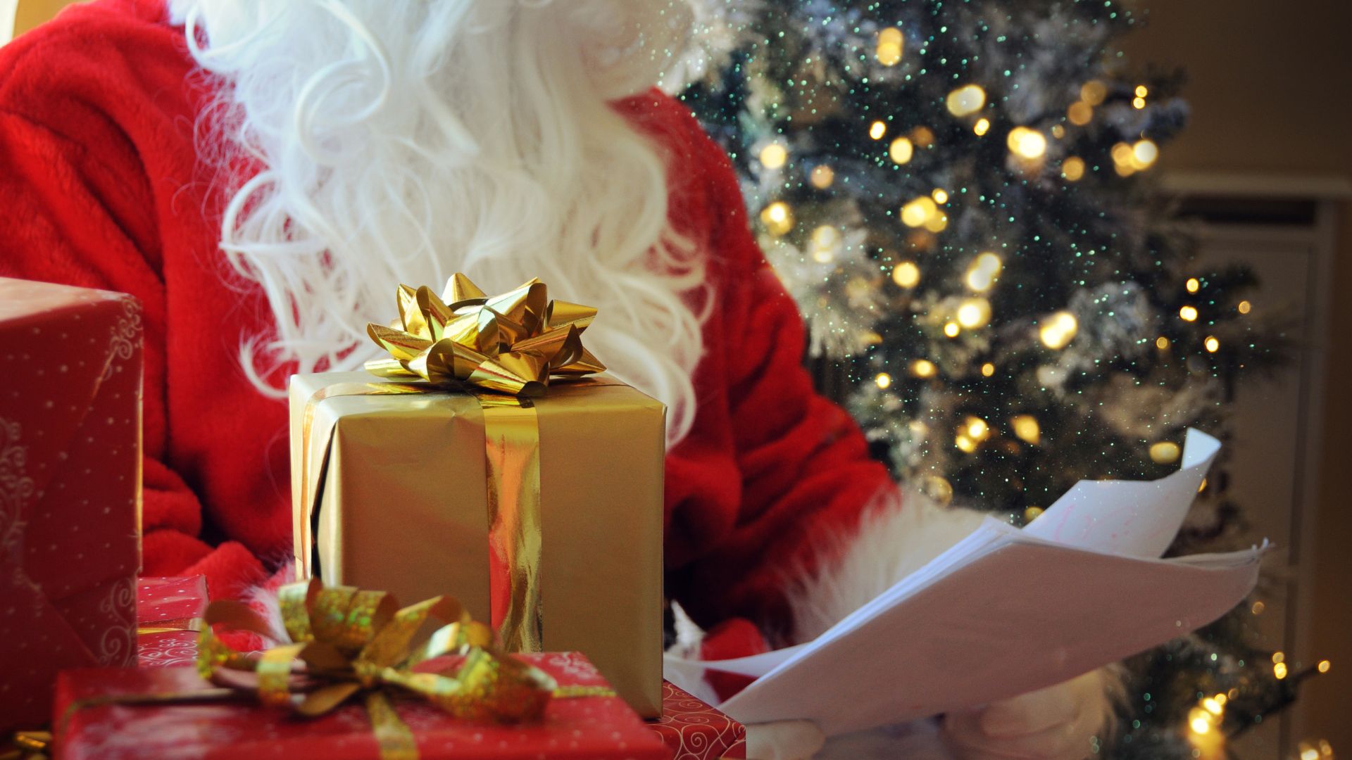 Santa Claus holding papers sits next to a decorated Christmas tree, with wrapped presents featuring gold bows in the foreground.