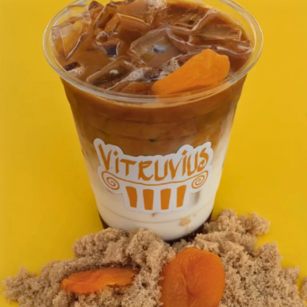 Iced coffee in a Vitruvius branded plastic cup, garnished with dried apricots and served with a pile of brown sugar on a yellow surface.