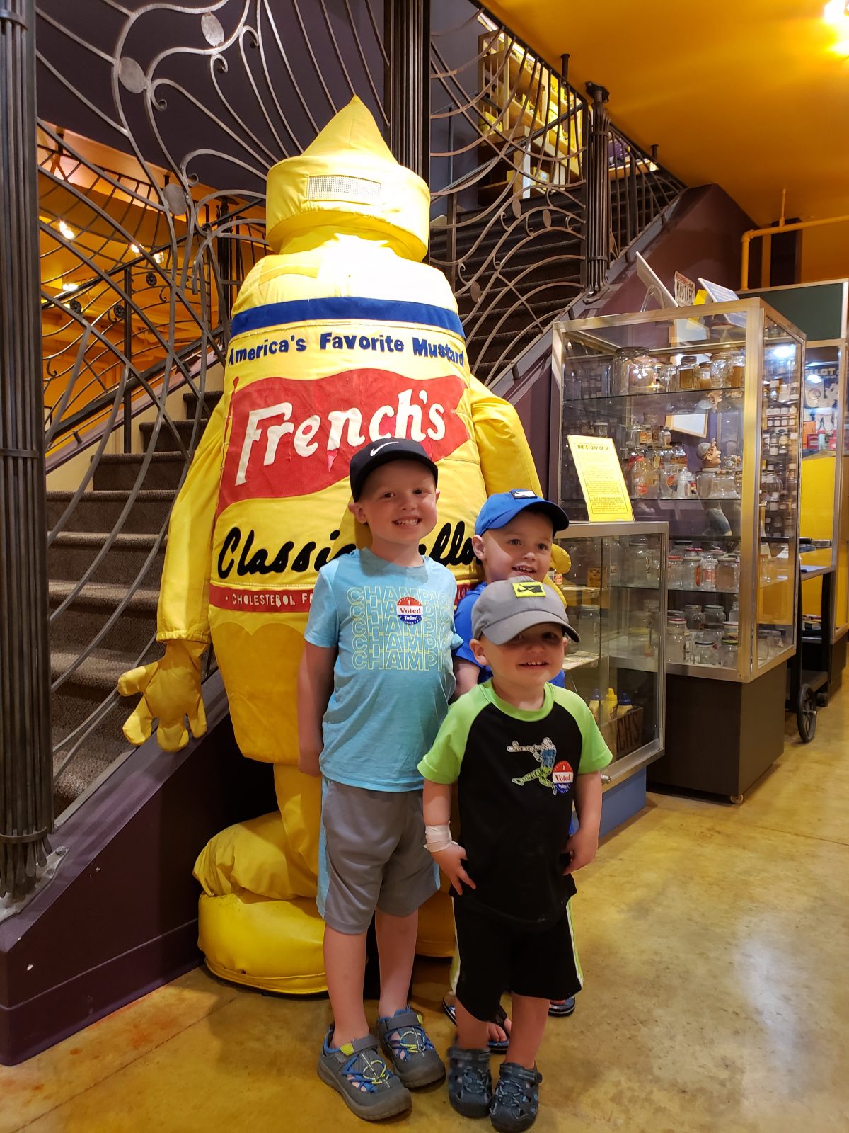 Three boys standing in front of a giant french's mustard statue.