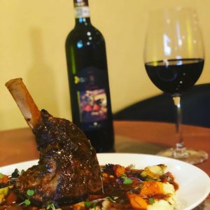 A lamb shank served over vegetables at an Easter brunch with friends, accompanied by a glass and bottle of red wine in the background.