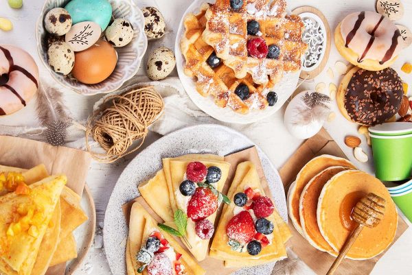 Assorted breakfast dishes including pancakes, waffles, and pastries, garnished with fresh berries and served with honey and eggs on a light background.