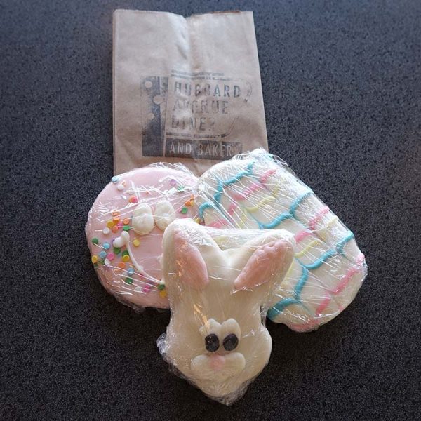A bunny-shaped and egg-shaped sugar cookies decorated with pastel icing, wrapped in plastic on a grey surface, next to a paper bakery bag.
