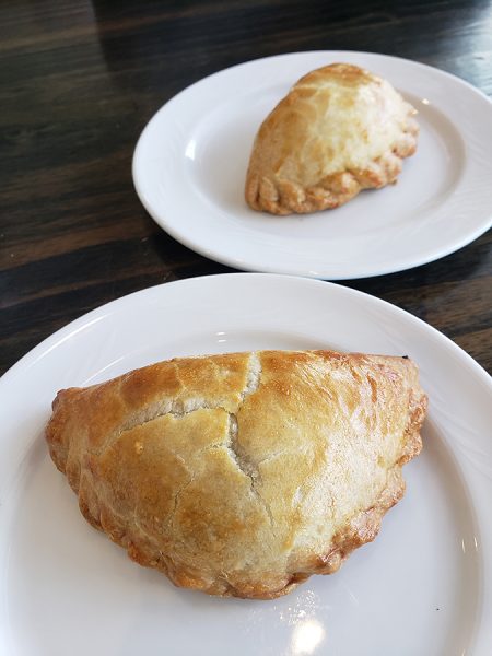Two pastries on white plates on a wooden table, one closer and larger, appearing as a golden-brown turnover, and the other smaller, possibly a sweet pastry.