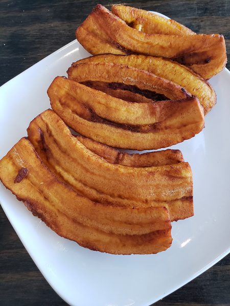 Fried plantain slices arranged in a curved pattern on a white square plate, showcased on a dark wooden table.