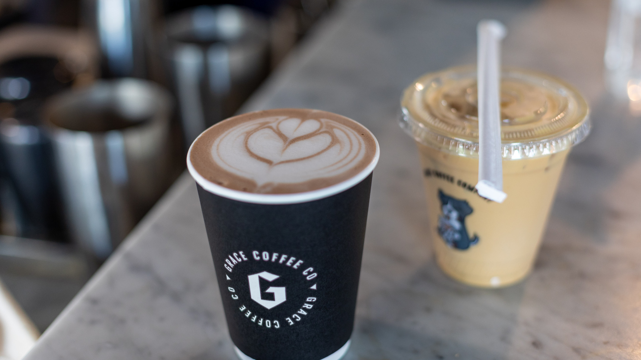 A paper cup cradles a hot coffee adorned with latte art, resting elegantly on a marble surface. Nearby, a plastic cup with a lid and straw holds a refreshing cold beverage—perfect springtime treats for enjoying the season's warmth.