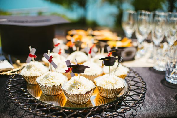 Assorted cupcakes with graduation cap toppings next to champagne glasses on a catering table.