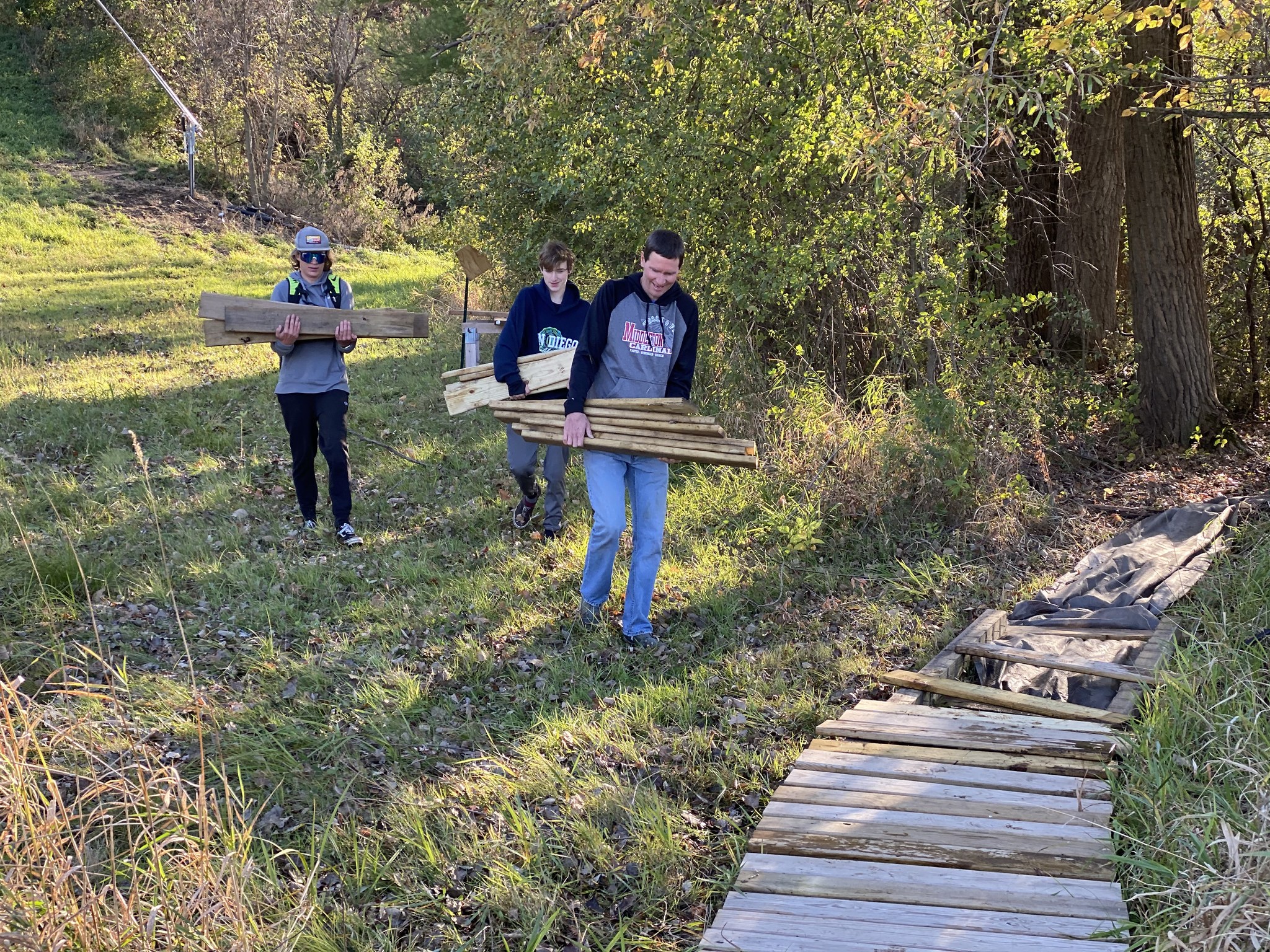 Three people carrying wooden planks on a grassy path next to a partially constructed wooden walkway in a wooded area.