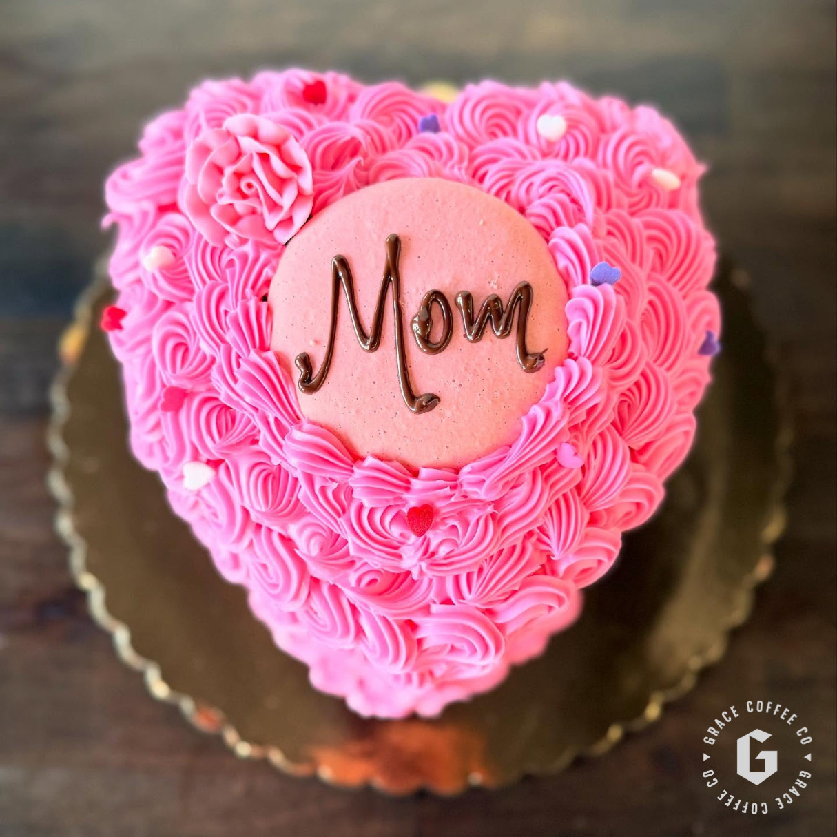 Heart-shaped pink frosted cake with a round plaque reading "Mom" in chocolate icing, decorated with piped rosettes and sprinkles on a gold cake board—perfect for Mother's Day treats or Mother's Day specials.