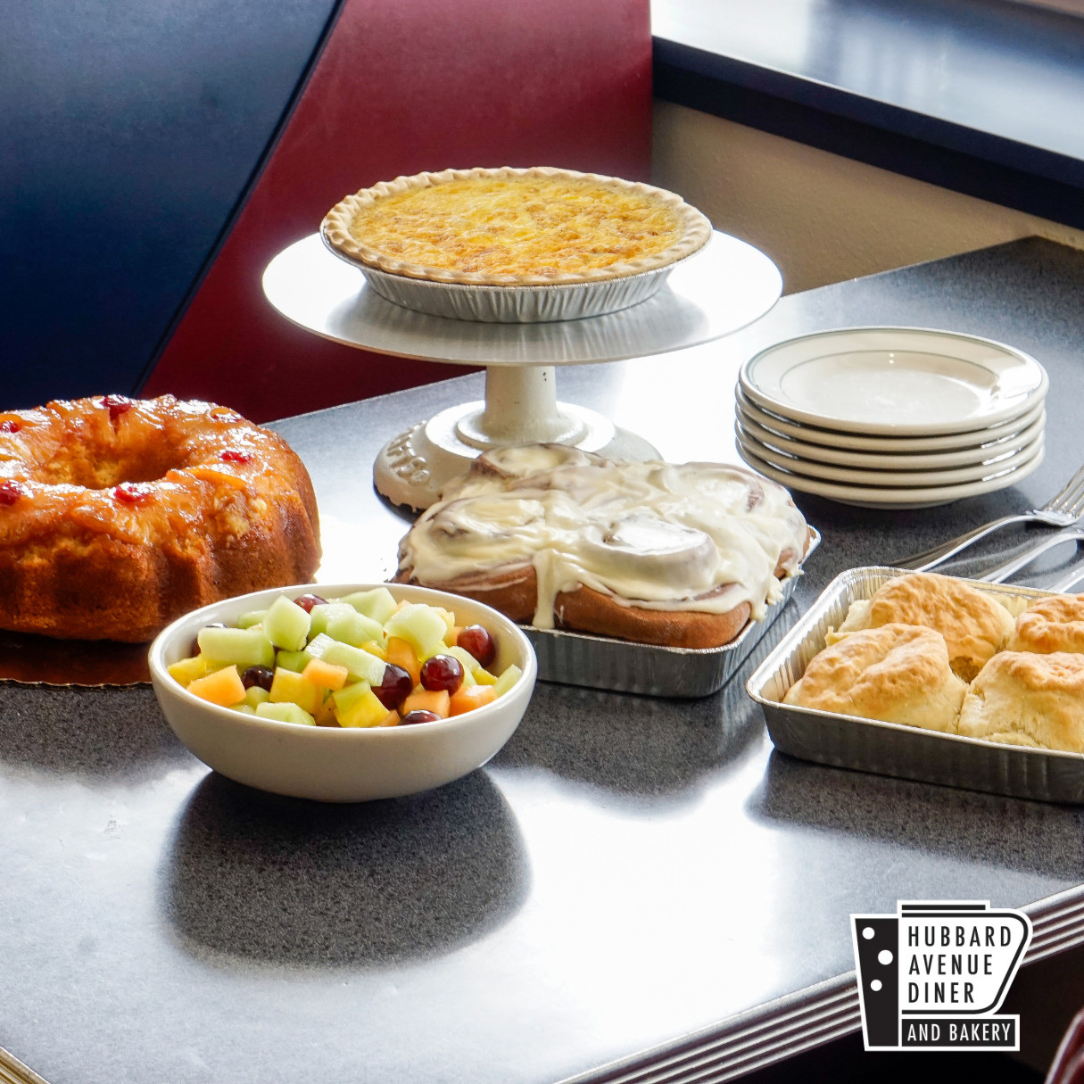 A table set for brunch with a bundt cake, fruit bowl, pie on a stand, iced cinnamon rolls, biscuits, and a stack of plates—perfect Mother's Day treats. Hubbard Avenue Diner and Bakery logo is in the corner.