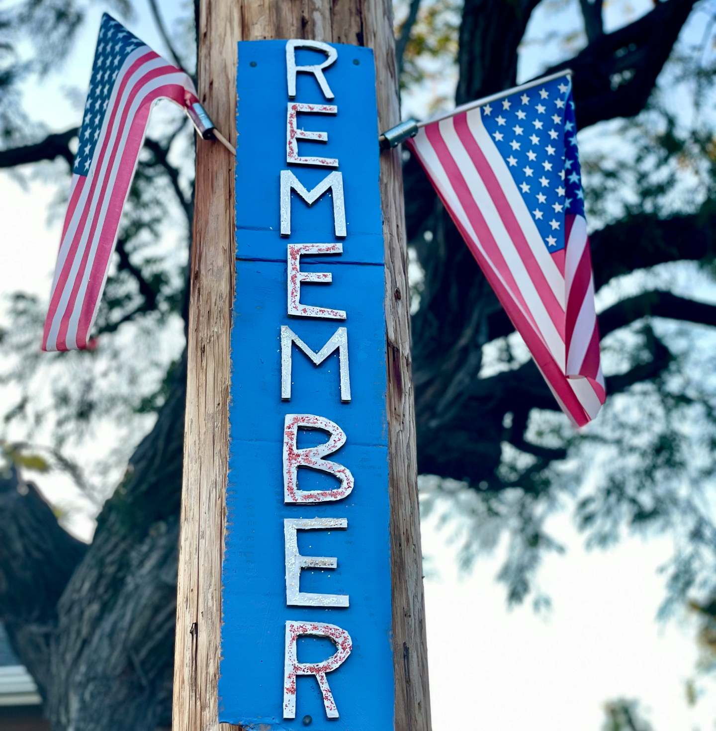 A vertical wooden post with the word "REMEMBER" in block letters and two American flags attached, one on each side, against a background of trees.