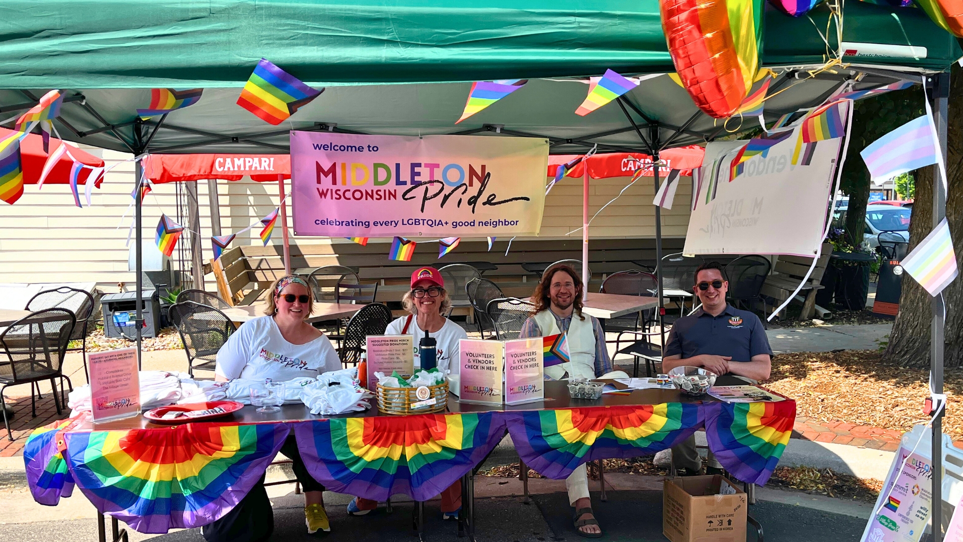 Four people sit at a decorated table under a tent at the Middleton Pride event in Wisconsin, surrounded by rainbow flags and banners promoting LGBTQIA+ inclusion.