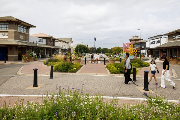 A suburban shopping village with people walking, benches, and a central fountain. The area is lined with a variety of stores and greenery.