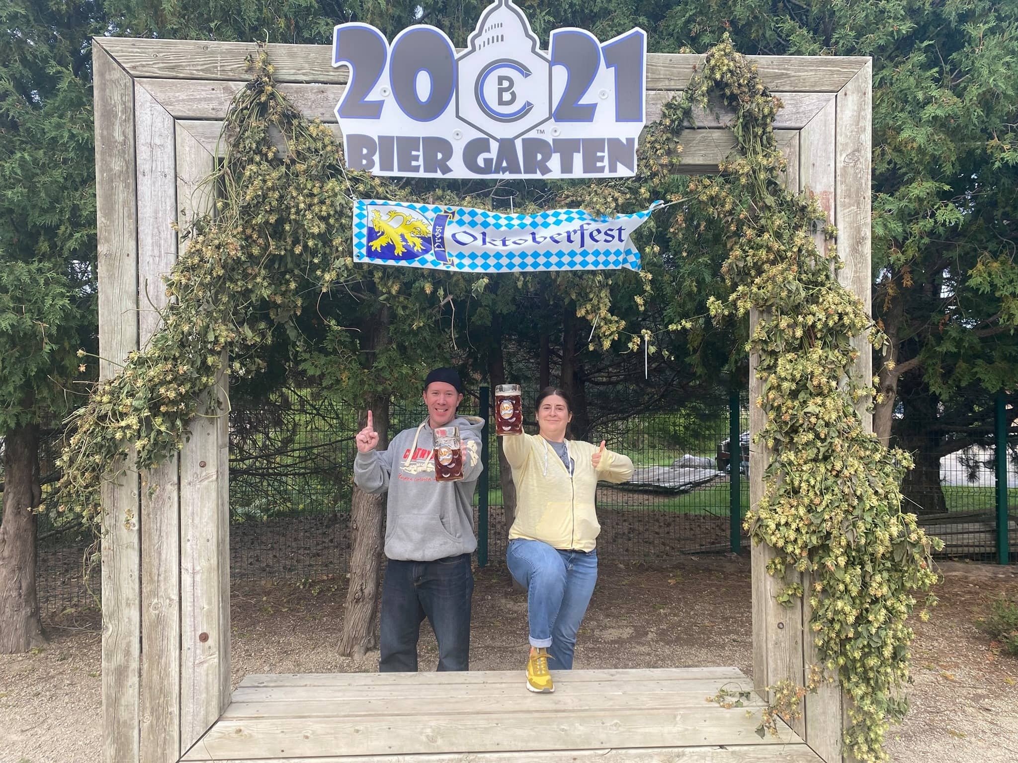 Two people pose under a 2021 Bier Garten sign framed with greenery, as an Oktoberfest banner hangs overhead, hinting at the lively atmosphere complete with live music.