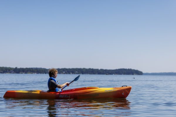 A person in a life jacket kayaks on calm water near Marshall Park, with a distant tree-lined shore under a clear blue sky—a perfect day to explore with Marshall Boats Board and Rentals.