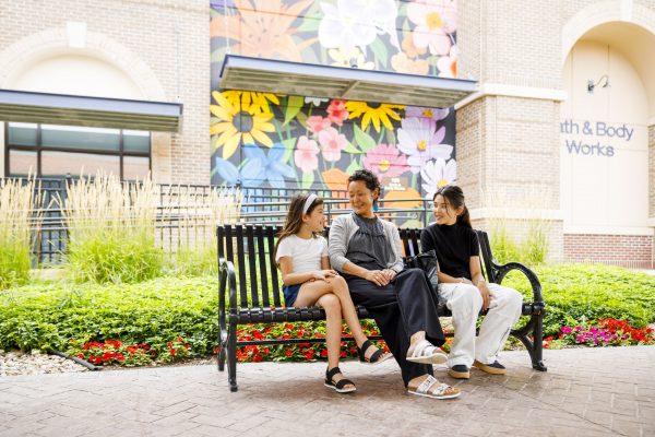 Three people are sitting on a black metal bench in front of a colorful floral mural and a "Bath & Body Works" store. They appear to be engaged in a friendly conversation.