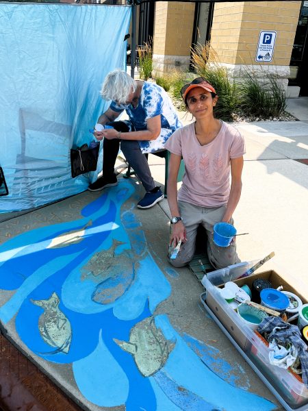 Two people work on a sidewalk chalk mural with blue water and fish outside a building; one paints while the other sits and mixes paint.