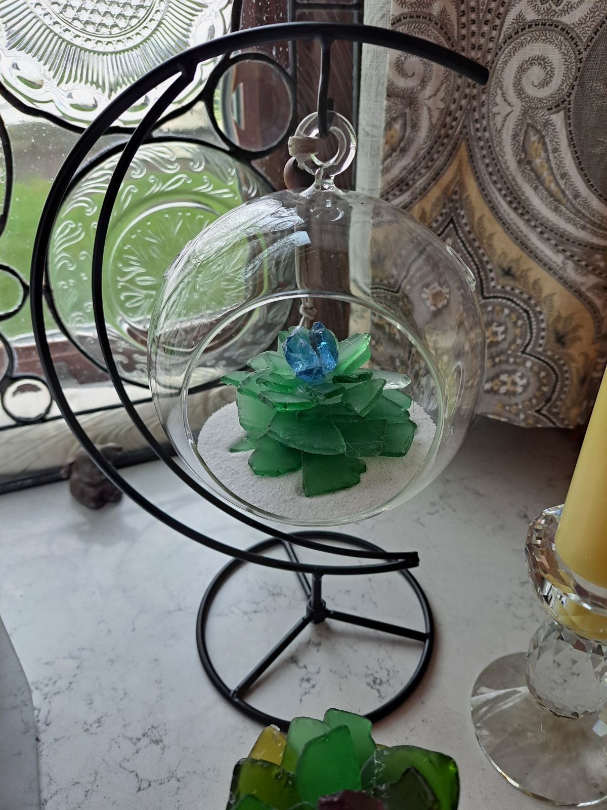 Glass terrarium with green glass pieces arranged as a succulent, featuring a blue glass piece on top, set on white sand, with a decorative background.