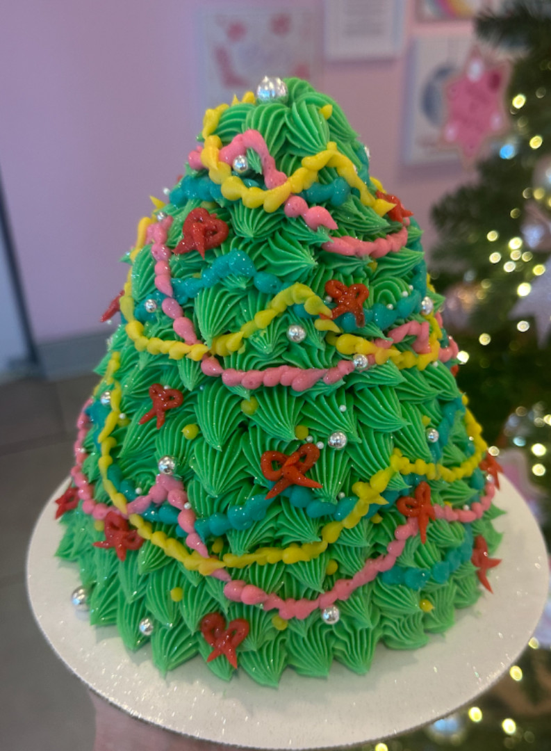 A Christmas tree-shaped cake with green frosting, decorated with pink and yellow icing, red bows, and silver balls, on a white base.