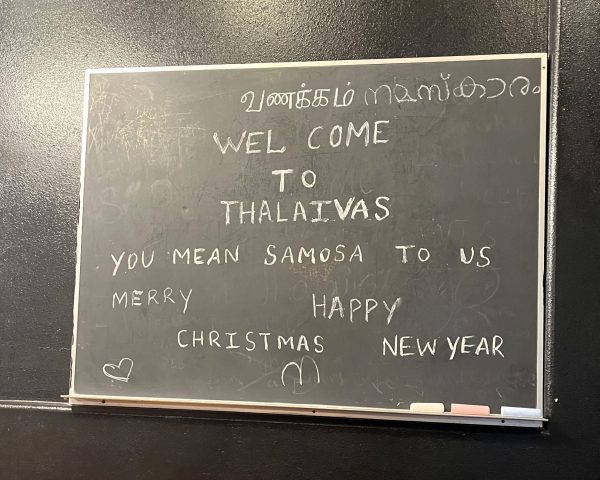 A chalkboard with messages in English and Tamil, reading: "WEL COME TO THALAIVAS. YOU MEAN SAMOSA TO US. MERRY CHRISTMAS HAPPY NEW YEAR" with heart and smiley face drawings.